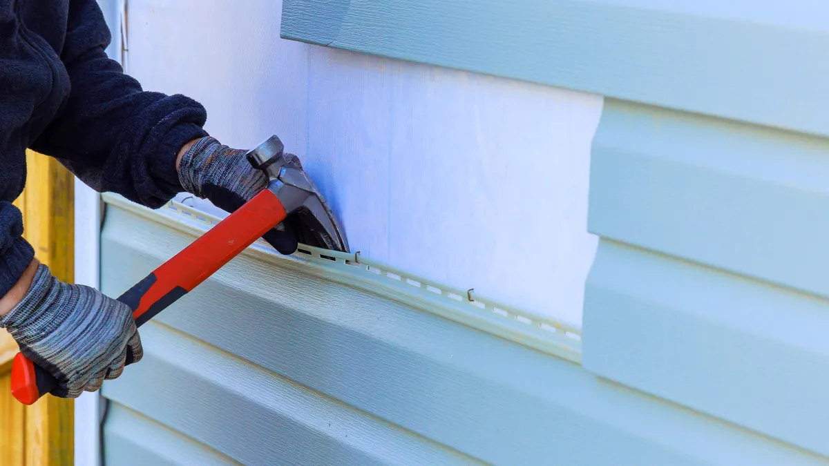 A gloved worker using a hammer to remove a damaged row of light blue vinyl siding from a house wall.