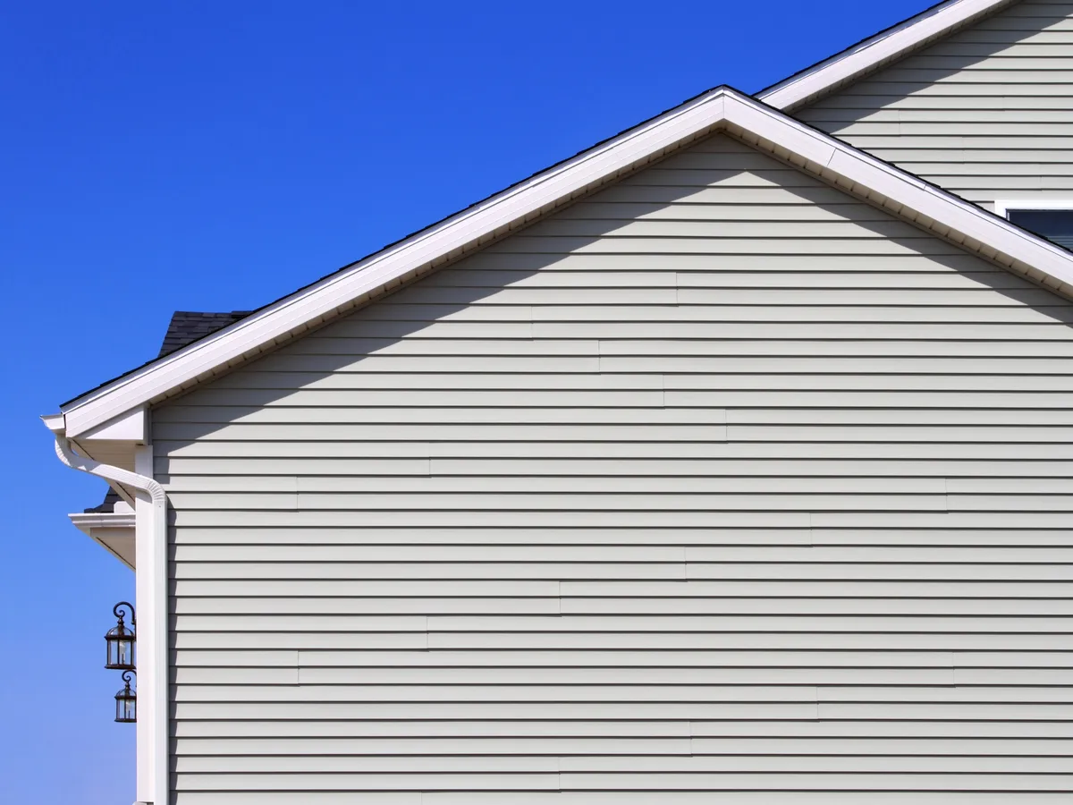 The gable end of a house with gray lap siding, white trim and gutters under a clear blue sky.